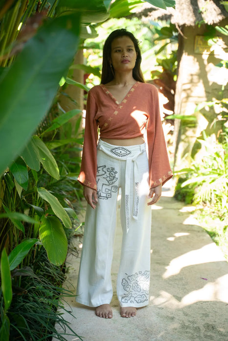 Woman in a rust-colored top and white pants with a pattern, standing in a lush green outdoor setting.