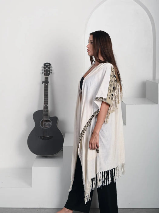 Woman wearing a white shawl with black patterns standing next to an acoustic guitar in a minimalistic setting.