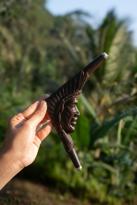 Hand holding a wooden smoking pipe with an engraved face against a blurred natural background
