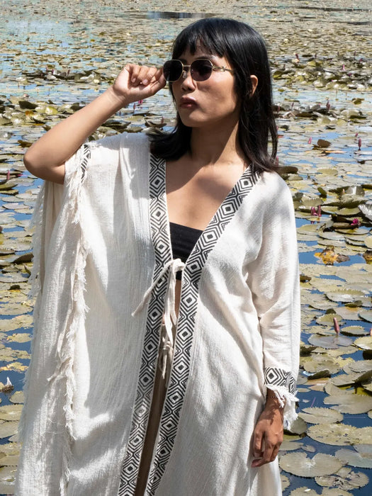 Woman wearing a white kimono-style garment with a geometric pattern, standing in front of a water lily pond.
