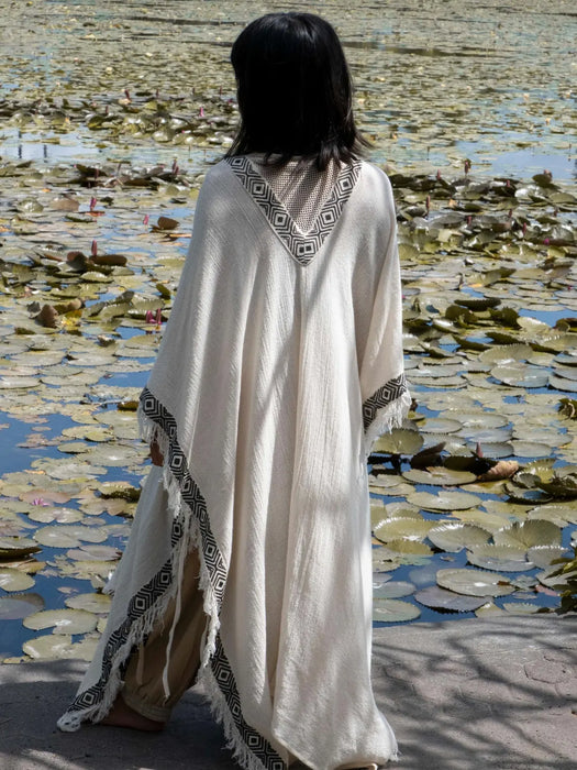 Person wearing a white dress with black patterns standing by a water body with lily pads.