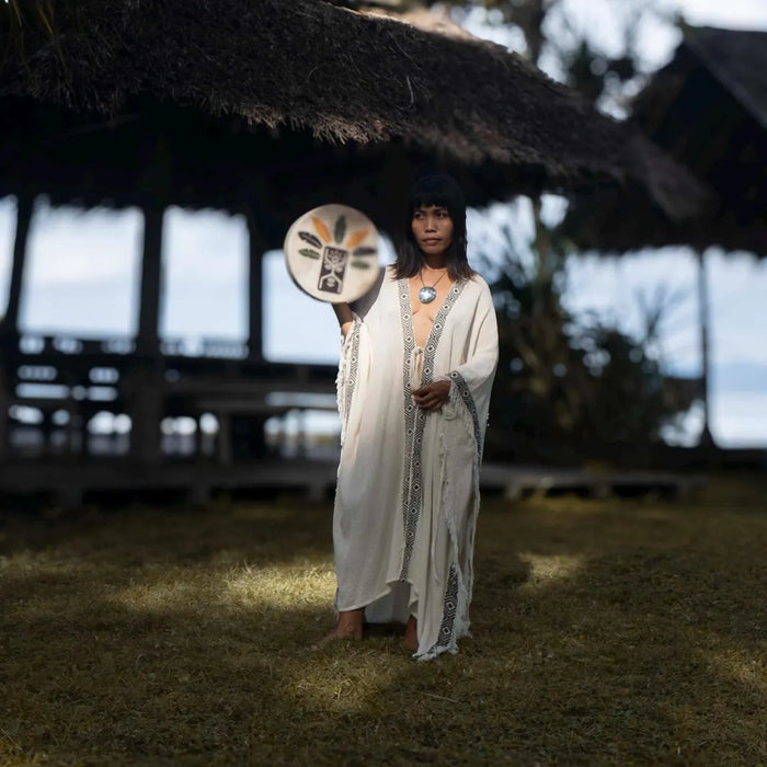 Person in traditional attire holding a ceremonial object in front of a thatched structure.