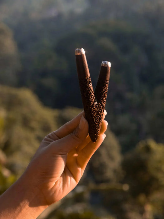 Hand holding two intricately designed wooden sticks against a blurred natural background