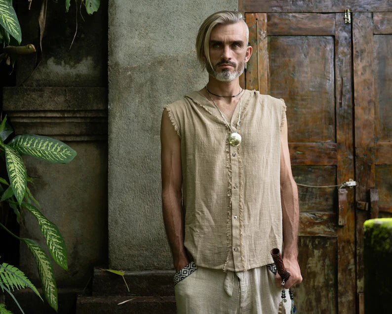 Man in a beige sleeveless shirt and white pants standing in front of a rustic wooden door.