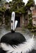 A polished silver metal object with ornate engraved detailing and a dark circular stone accent, balanced on a black surface with a white feather in the foreground and a mossy stone doorway behind it.