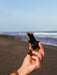 Hand holding a small black device with gold accents on a beach