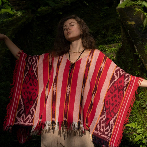 A woman extends her arms amidst lush greenery, wearing a short, handmade Boho poncho, epitomizing the free-spirited essence of shamanic and hippie clothing